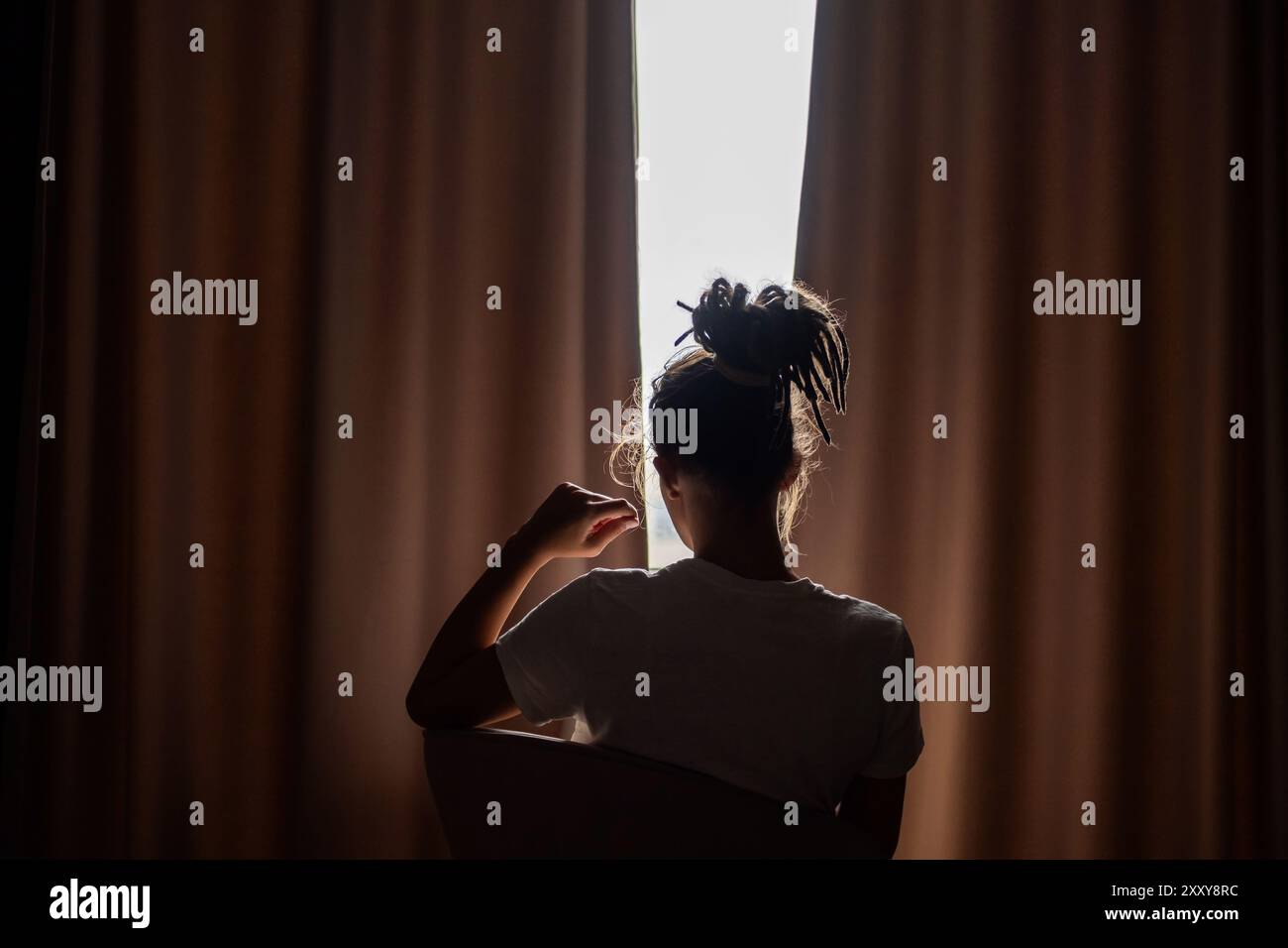 Silhouette de la personne assise contre la lumière tout en regardant à travers les rideaux. Banque D'Images