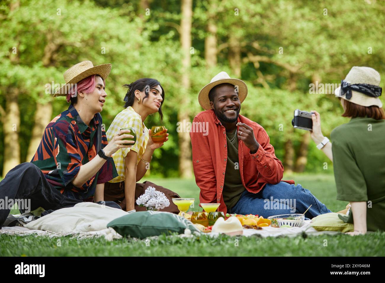 Groupe d'amis assis sur une couverture de pique-nique dans un parc verdoyant luxuriant, partageant des boissons et des collations, une personne capturant des souvenirs avec un smartphone tandis que d'autres s'engagent dans une conversation joyeuse Banque D'Images