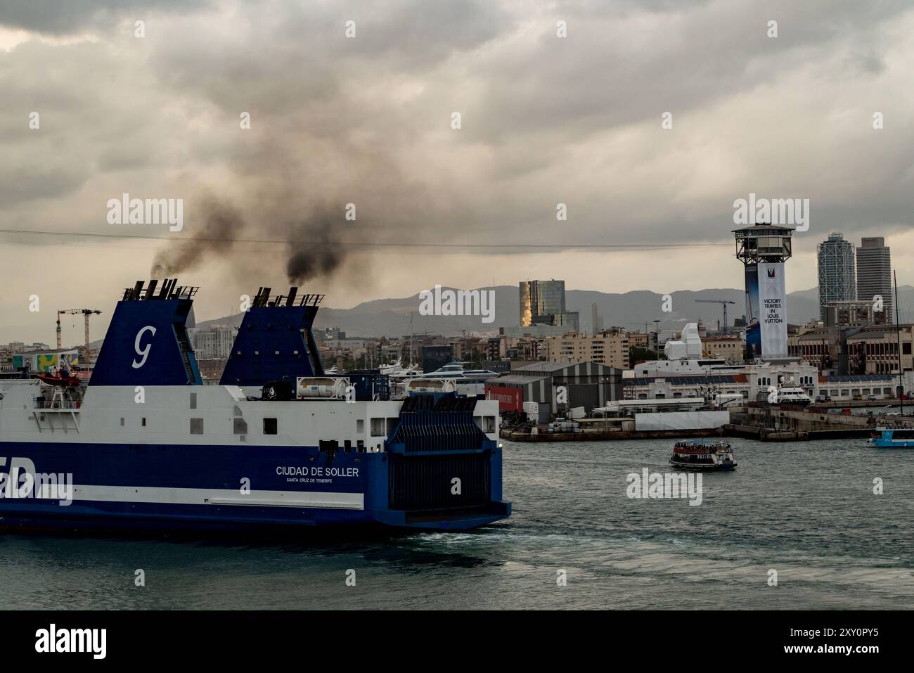 Dans le port de Barcelone, les entonnoirs d’un ferry dégagent de la fumée lorsqu’il passe devant la tour Sant Sebastia décorée de la marque Louis Vuitton en tant que sponsor de la 37ème Coupe de l’America, compétition de voile organisée dans la capitale catalane. Les émissions de dioxyde de soufre et d'oxyde d'azote rendent le port de Barcelone pire en Europe pour la pollution atmosphérique, principalement pour les navires de croisière. Banque D'Images