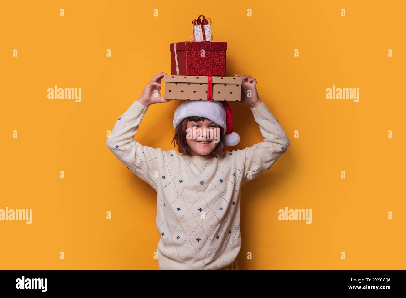 Une jeune fille preteen joyeuse portant un chapeau de Père Noël, équilibrant une pile de cadeaux de Noël sur sa tête, souriant vigoureusement sur un fond jaune vif. Banque D'Images