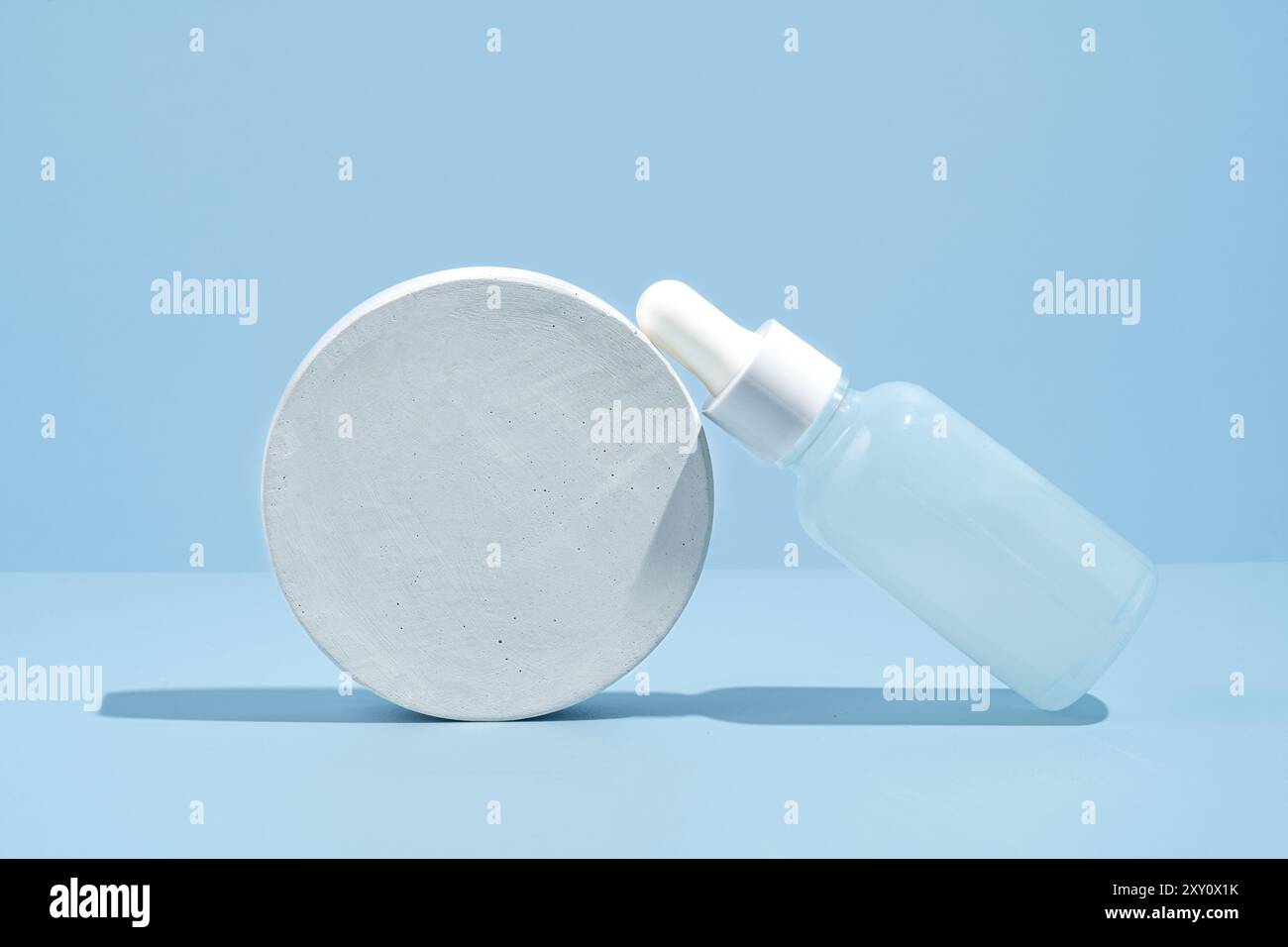 Un flacon compte-gouttes de sérum en verre dépoli repose à côté d'une plate-forme ronde en pierre blanche sur un fond bleu pastel créant une scène cosmétique sereine. Banque D'Images