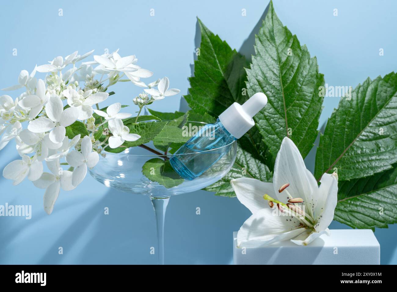 Un sérum hydratant pour le visage dans un compte-gouttes en verre se trouve élégamment sur un verre à cocktail, entouré d'hortensia blanches et de lis sur un fond bleu apaisant, SY Banque D'Images