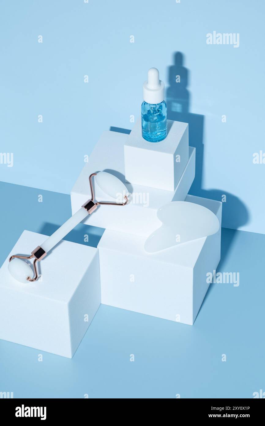 Un arrangement serein d'outils de soin de la peau et une bouteille de sérum hyaluronique sur des blocs géométriques, projetant des ombres douces sur un fond bleu. Ajoute un luxe Banque D'Images