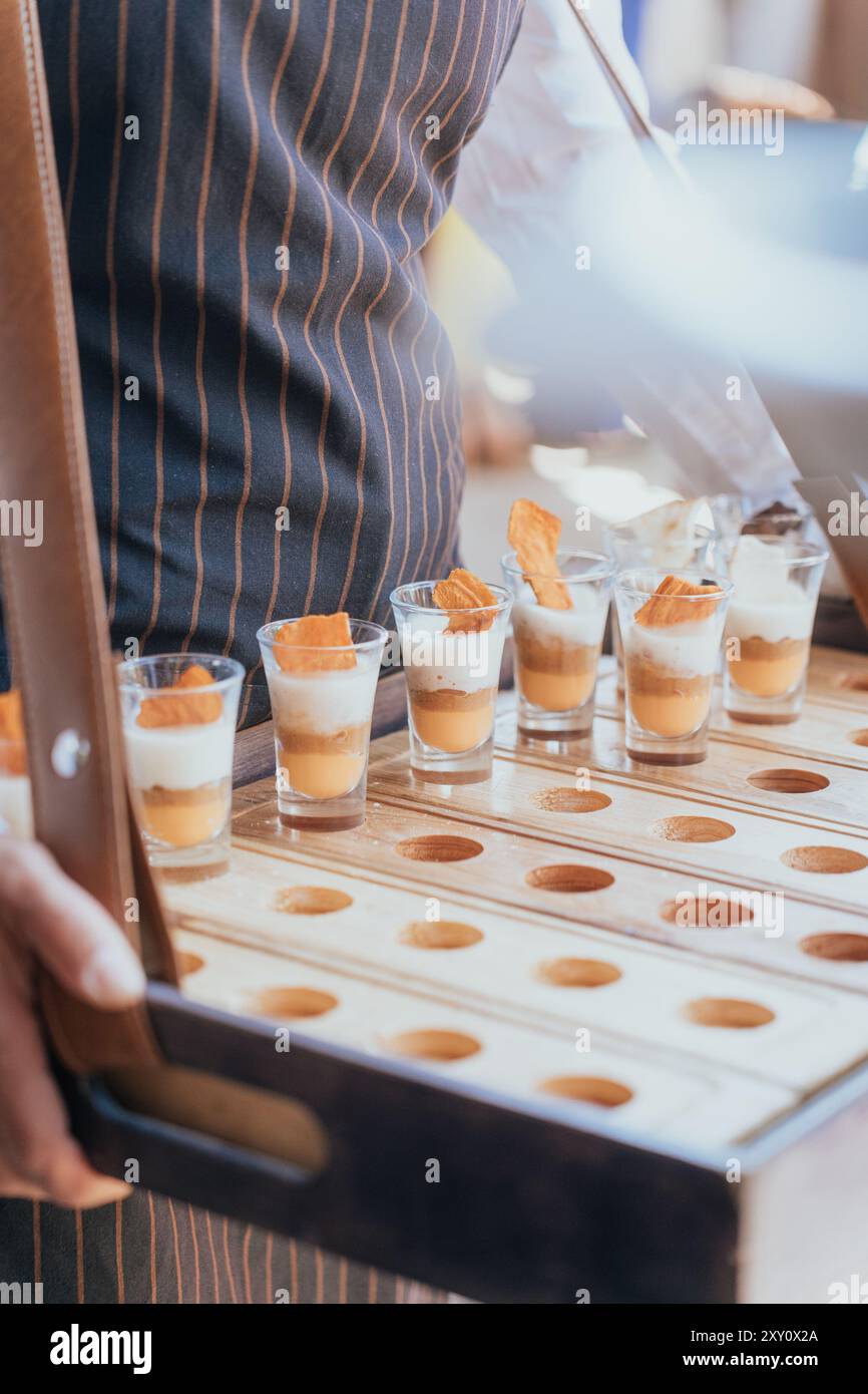 Un serveur dans un tablier rayé présente un plateau en bois avec des desserts gourmands lors d'un événement de mariage le plateau est conçu avec des découpes circulaires, chacune tenant un Banque D'Images