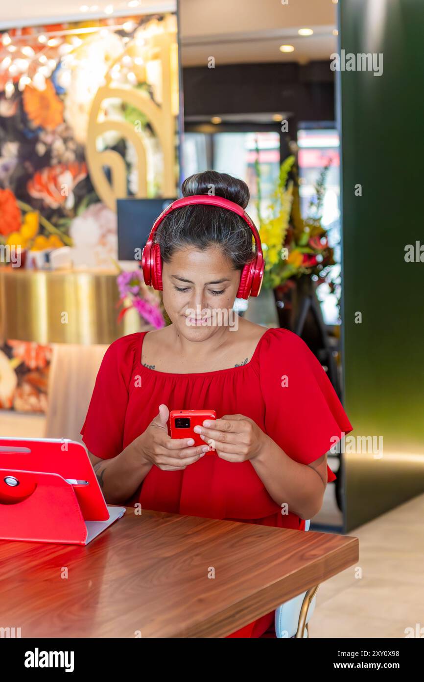 Une femme vêtue d'une robe rouge éclatante et d'écouteurs utilise son smartphone, souriant tout en profitant éventuellement d'un contenu attrayant dans un environnement de travail où Se trouve Une tablette Banque D'Images