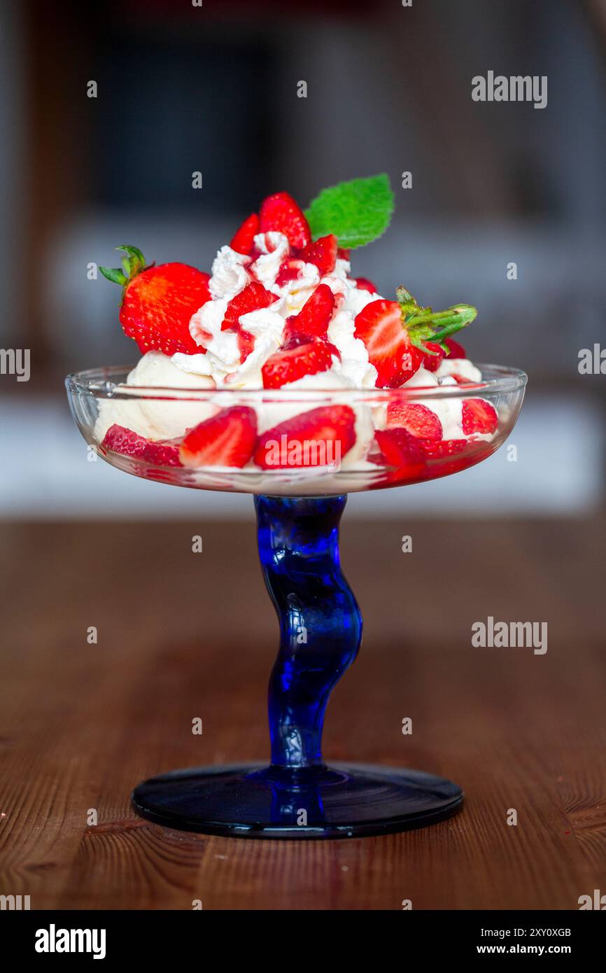 Une délicieuse portion de crème glacée à la vanille garnie de fraises fraîches tranchées et de crème fouettée, présentée dans un élégant bol en verre à tige bleu. Banque D'Images