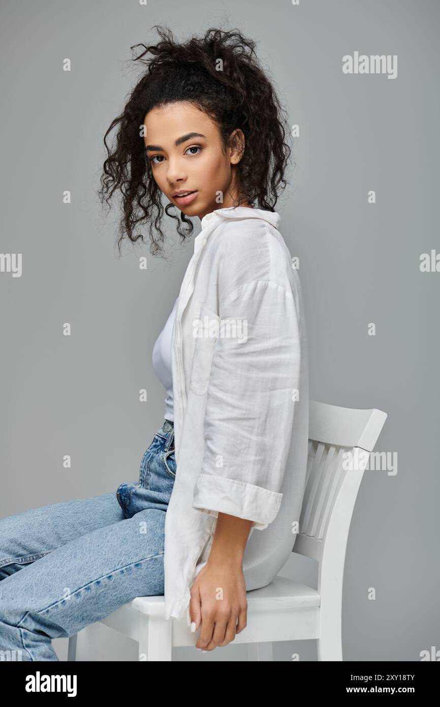 Une jeune femme afro-américaine est assise en toute confiance sur une chaise blanche sur un fond gris, portant une chemise blanche et un Jean bleu. Banque D'Images