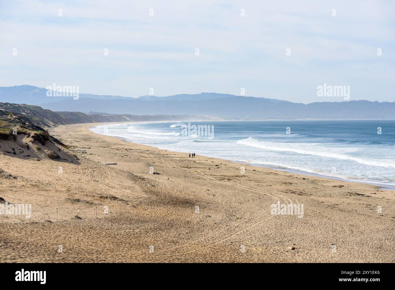 Les gens marchant le long d'une grande plage de sable bordée de hautes dunes de sable un jour d'automne brumeux. D'énormes vagues se brisant sur la plage sont visibles à distance. M Banque D'Images