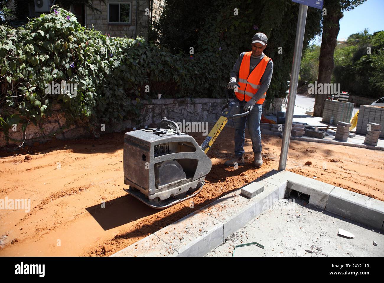 Un travailleur palestinien avec gilet de protection utilise un compacteur Bomag avec plaque vibrante pour comprimer le sol de sable rouge sur un trottoir où les briques seront posées Banque D'Images Un travailleur palestinien avec gilet de protection utilise un compacteur Bomag avec plaque vibrante pour comprimer le sol de sable rouge sur un trottoir où les briques seront posées Banque D'Images