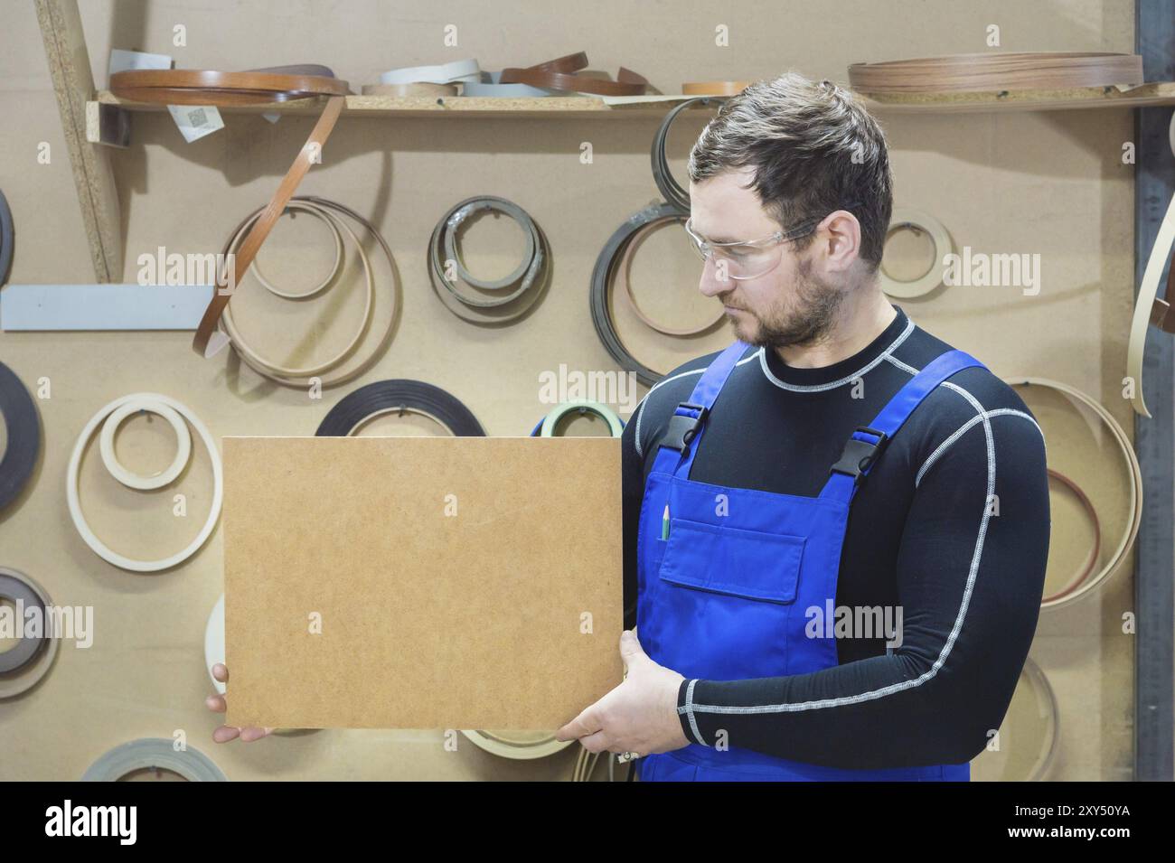 Le fabricant de meubles dans des vêtements spéciaux et des lunettes garde une plaque vide pour le texte sur leur lieu de travail. Fabrication artisanale de meubles Banque D'Images