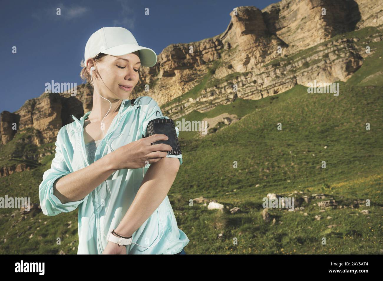 Une femme sportive dans une casquette et nuzhniki commute les chansons sur son téléphone portable lors d'un exercice en plein air sur fond de champs et de rochers Banque D'Images Une femme sportive dans une casquette et nuzhniki commute les chansons sur son téléphone portable lors d'un exercice en plein air sur fond de champs et de rochers Banque D'Images