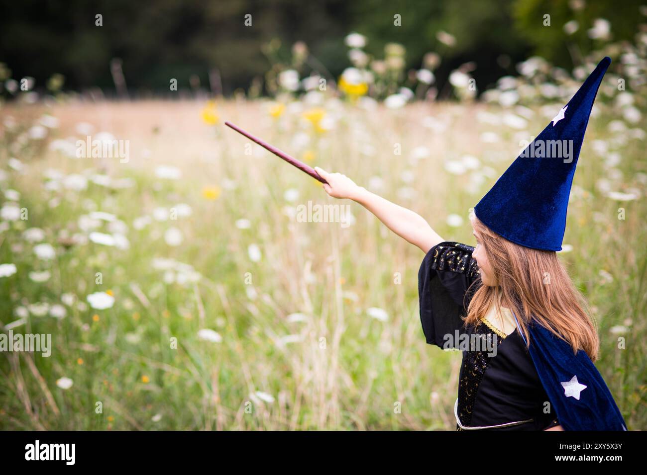 Fille d'enfant blonde caucasienne en costume bleu sorcier avec cape et chapeau, jetant un sort avec baguette magique dans le fond de champ extérieur. Banque D'Images