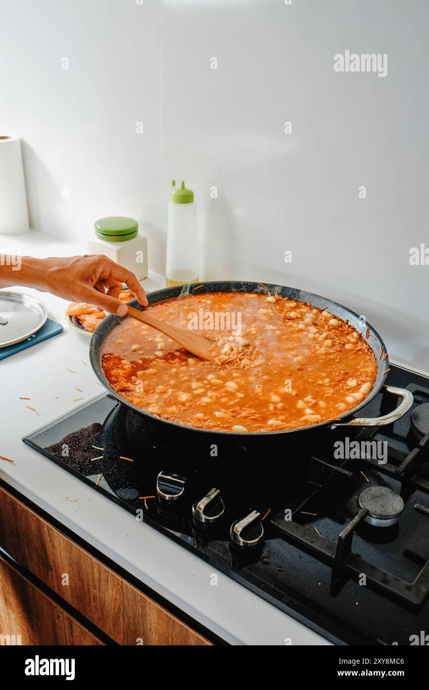 un homme remue la poêle à paella où il cuisine une fideua espagnole, dans le poêle d'une cuisine domestique Banque D'Images