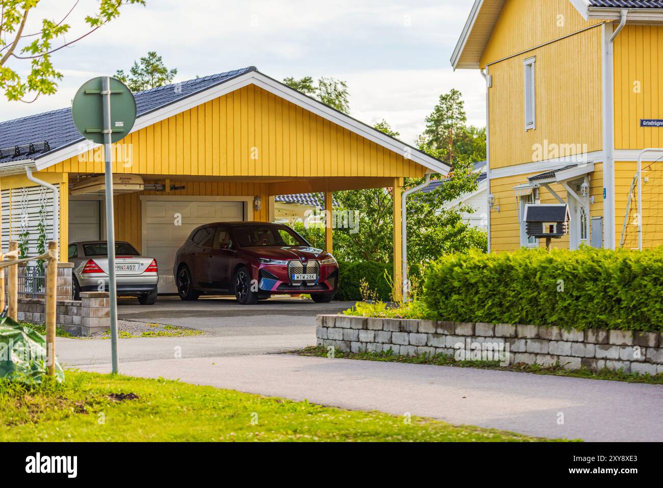 Gros plan de la villa avec maison en bois peint en jaune et garage, avec voiture électrique BMW iX40 et Mercedes à l'intérieur du garage. Banque D'Images