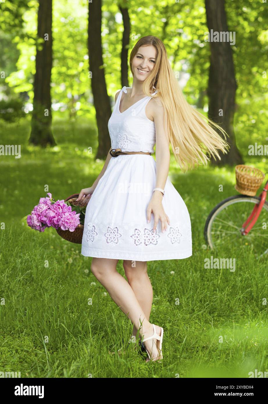 Belle femme blonde portant une belle robe s'amusant dans le parc avec vélo portant un beau panier plein de fleurs de pivoine. Paysage vintage. Joli Banque D'Images