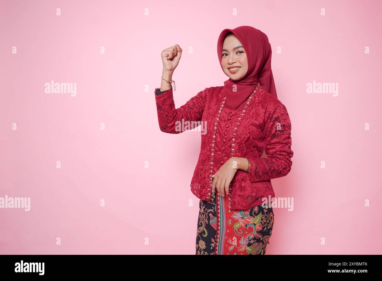 Femme asiatique joyeuse portant une robe kebaya moderne et hijab soulevant des mains de poing serrées geste d'indépendance isolé sur fond rose. Banque D'Images