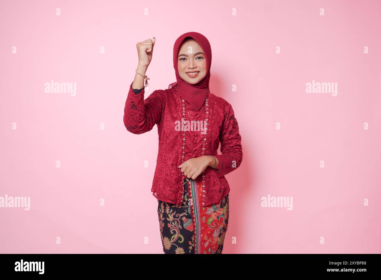 Femme asiatique joyeuse portant une robe kebaya moderne et hijab soulevant des mains de poing serrées geste d'indépendance isolé sur fond rose. Banque D'Images
