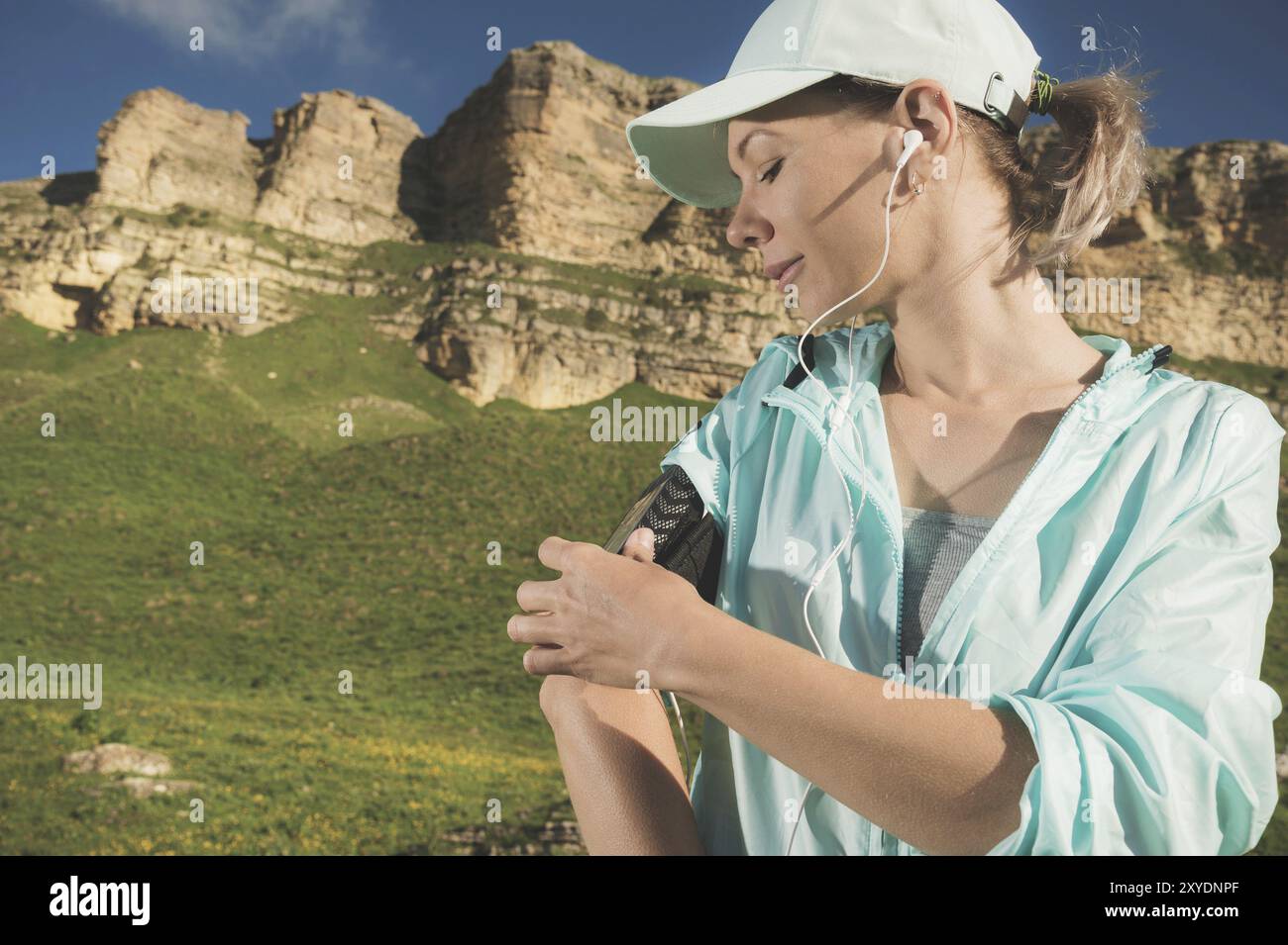 Une femme sportive dans une casquette et nuzhniki commute les chansons sur son téléphone portable lors d'un exercice en plein air sur fond de champs et de rochers Banque D'Images Une femme sportive dans une casquette et nuzhniki commute les chansons sur son téléphone portable lors d'un exercice en plein air sur fond de champs et de rochers Banque D'Images