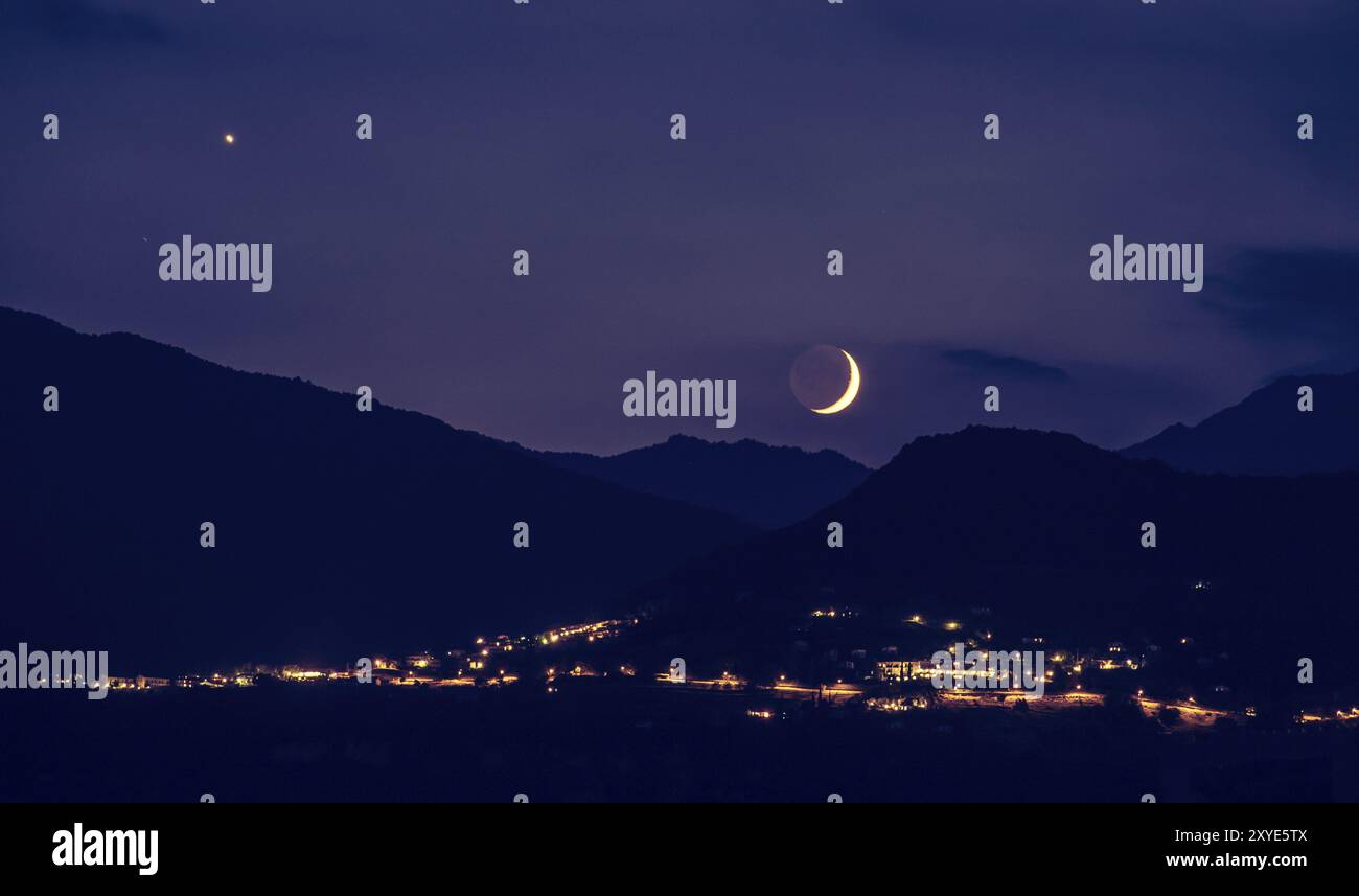 La demi-lune se lève sur un horizon pourpre, les montagnes et les lumières des villages Banque D'Images