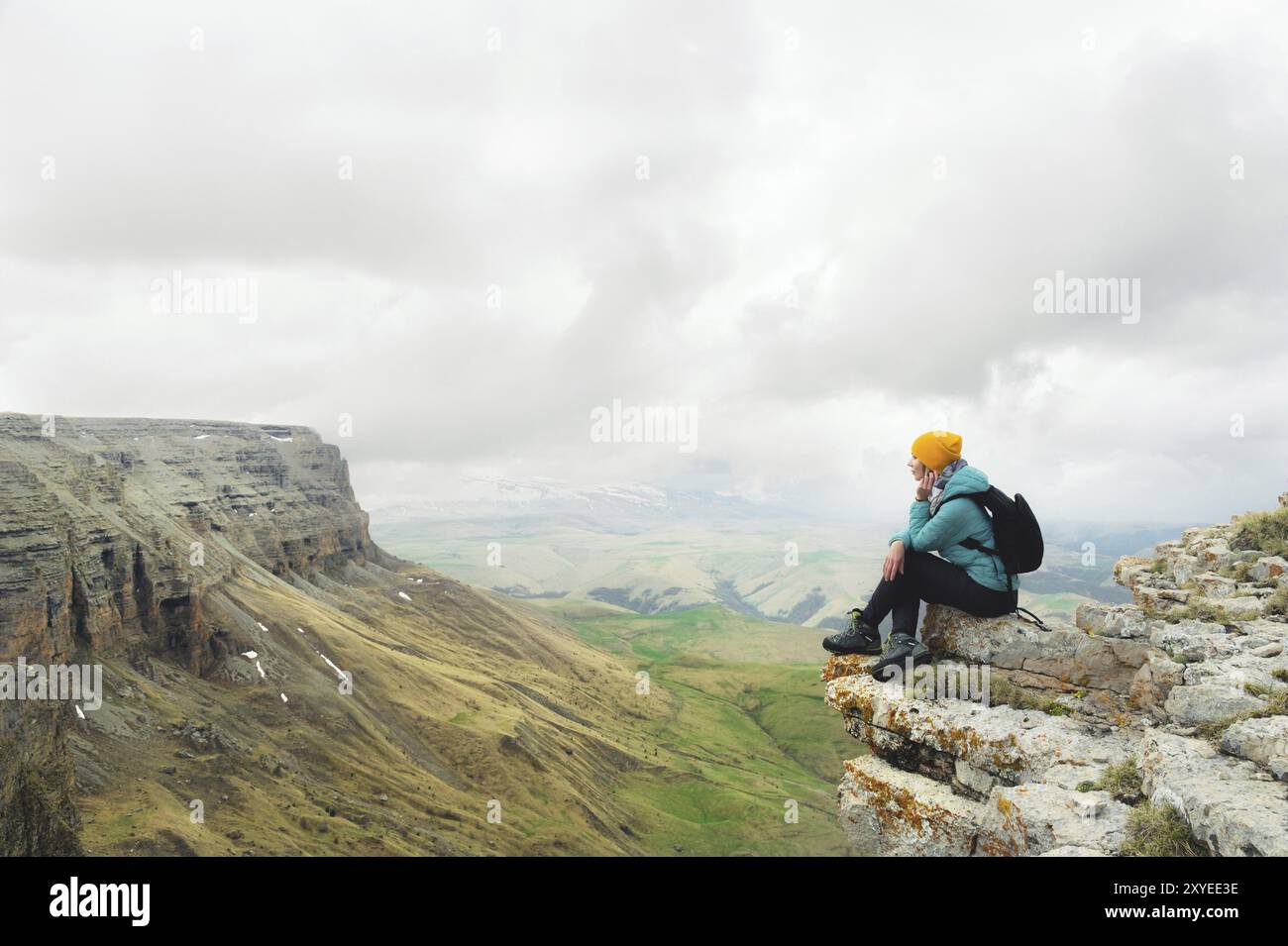 Jeune femme avec un sac à dos pensivement assis sur le bord d'un rocher et regardant le ciel avec des nuages Banque D'Images