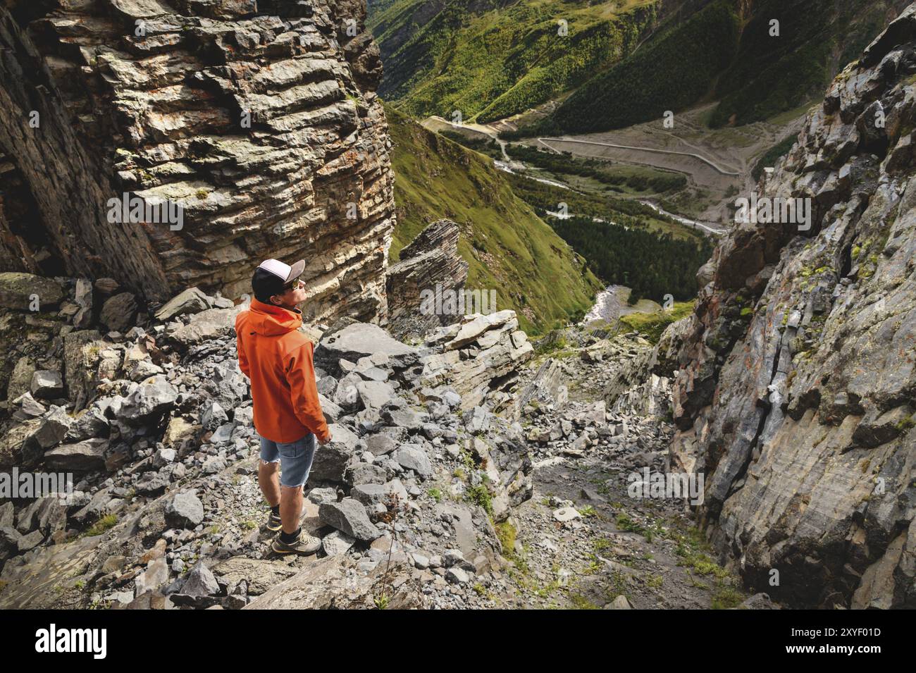 Portrait d'un randonneur masculin en short et une casquette à l'arrière se tient sur de hauts rochers au bord d'une falaise sur le fond d'une vallée verte W. Banque D'Images