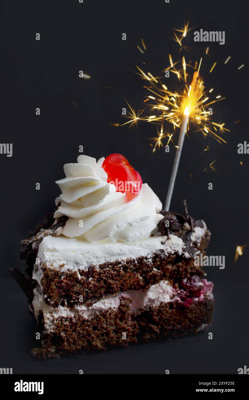 Un morceau de gâteau avec un bonbon rouge vif sur le dessus et un feu de Bengale brûlant sur un fond gris foncé Banque D'Images