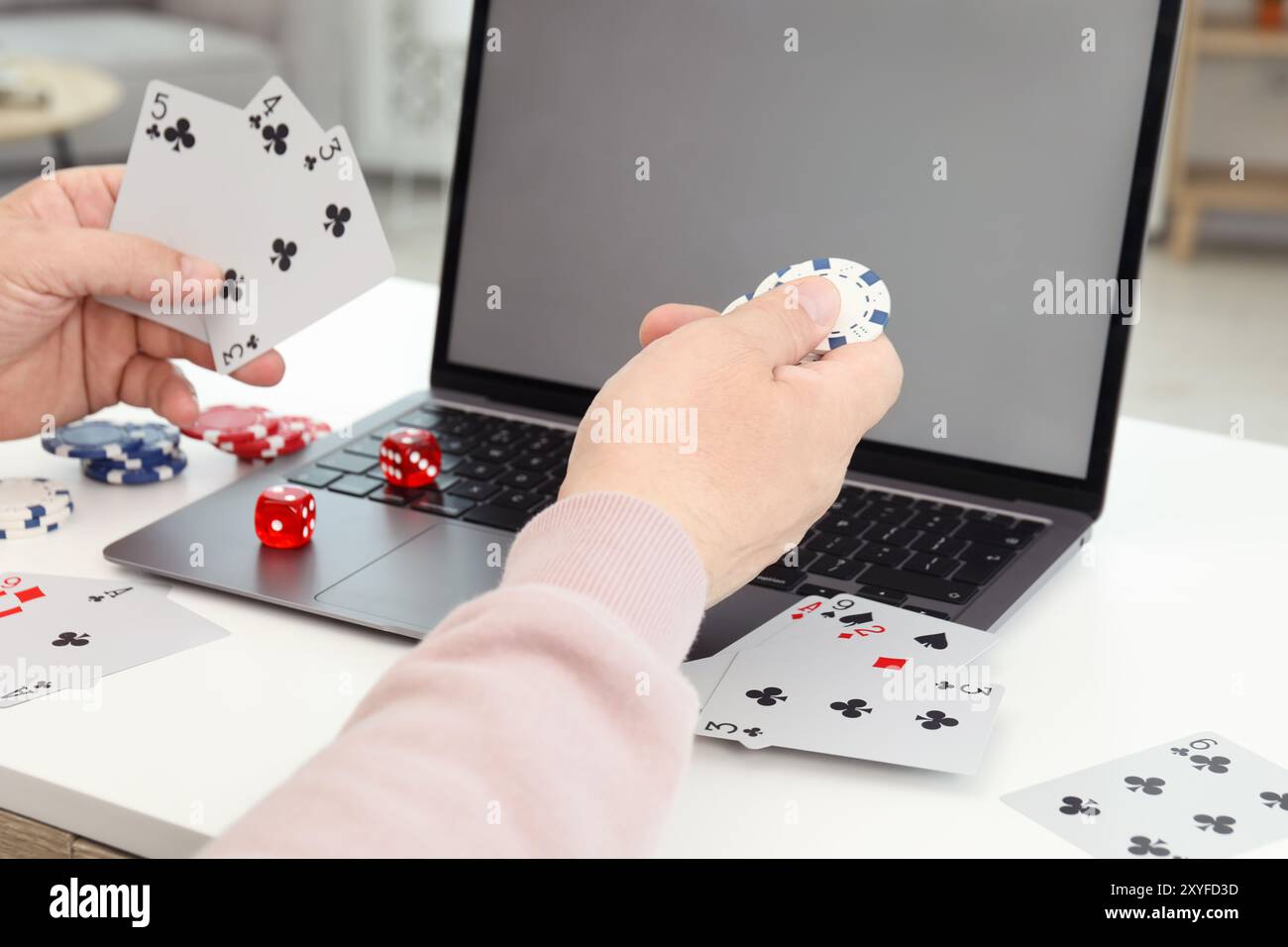 Poker en ligne. Homme tenant des cartes à jouer et jeton à la table blanche avec ordinateur portable, gros plan Banque D'Images