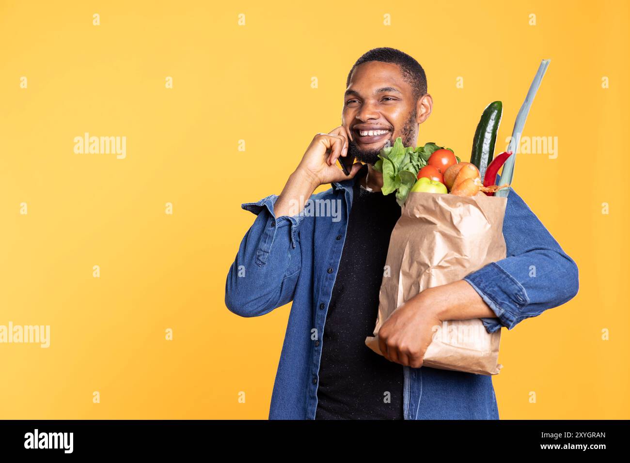 Homme afro-américain parle par téléphone avec des amis et tient un sac d'épicerie, magasinant pour des aliments bio frais et discutant avec des gens sur smartphone. Jeune homme avec des fruits et des légumes. Banque D'Images