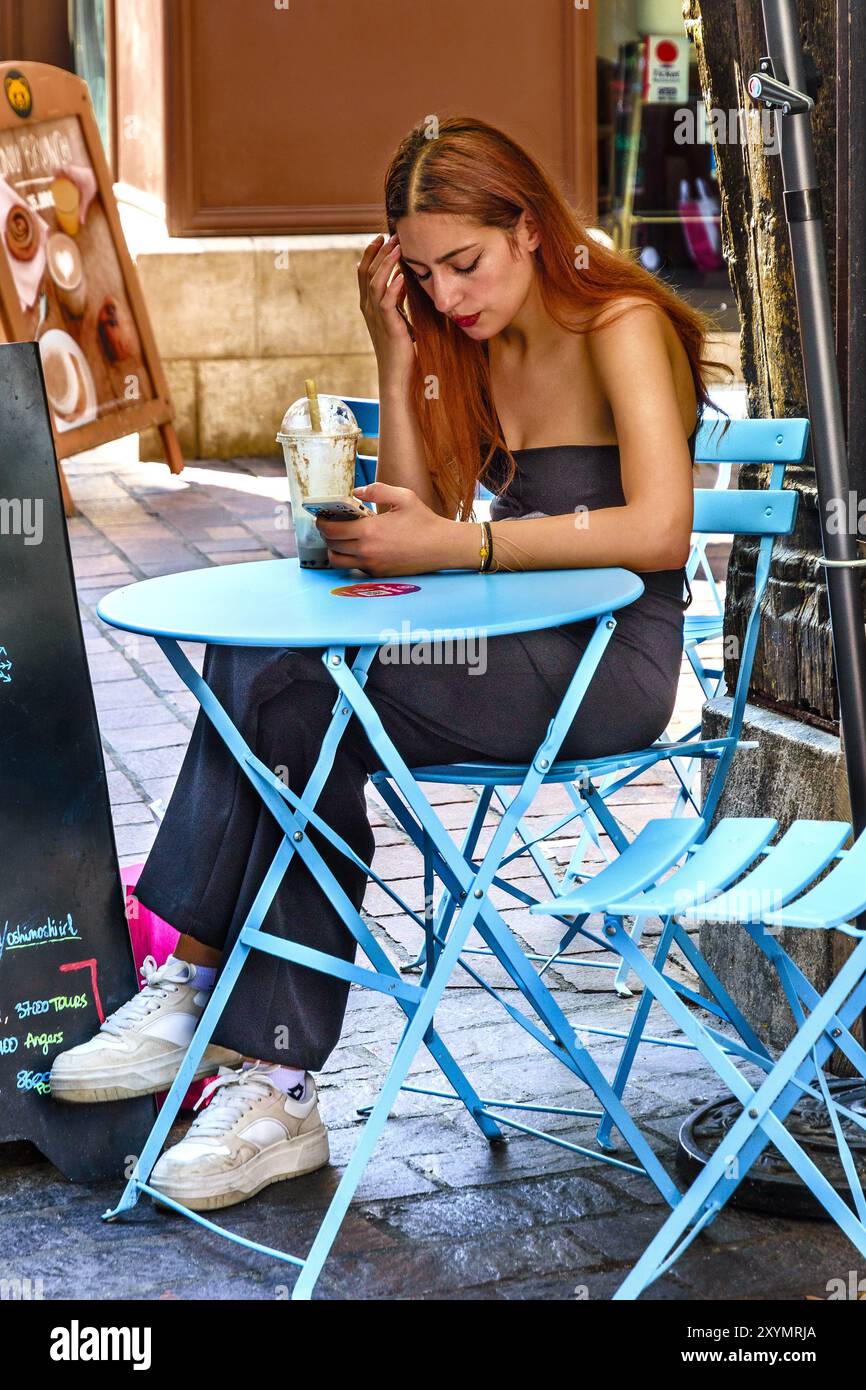 Jeune femme attrayante avec de longs cheveux auburn assise à la table du café regardant smartphone - Tours, Indre-et-Loire (37), France. Banque D'Images