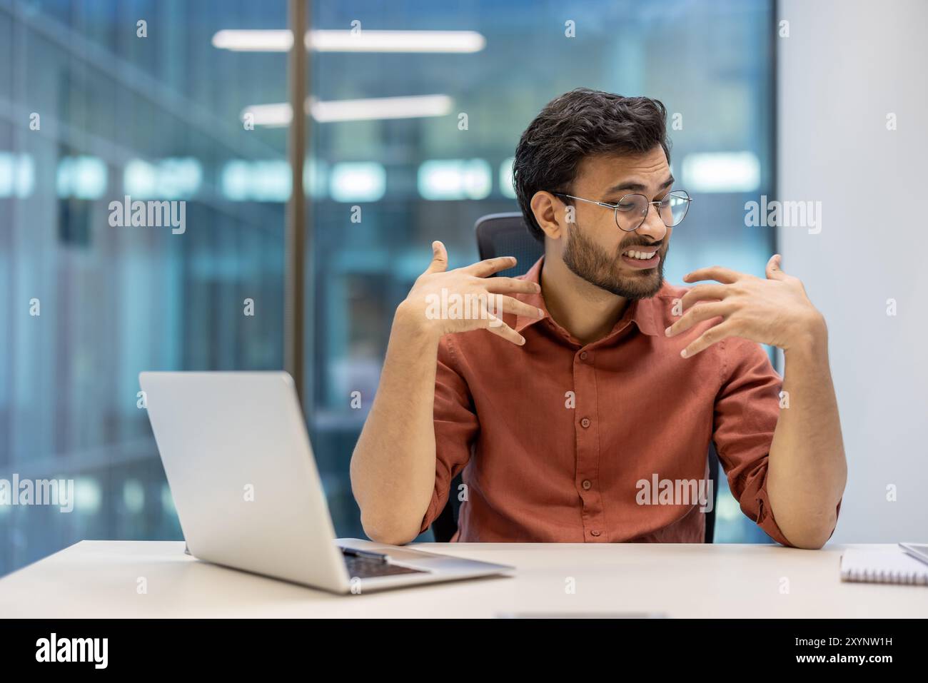 Les professionnels se sentent inconfortables en raison de la chaleur dans le bureau. Jeune homme faisant des gestes de frustration tout en travaillant au bureau avec un ordinateur portable. Surchauffe du lieu de travail causant du stress et des difficultés de communication. Banque D'Images