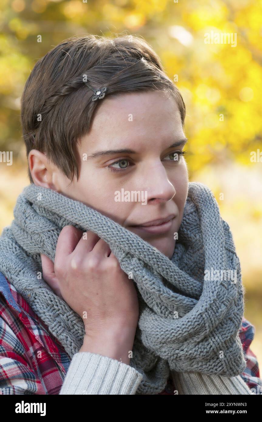 Jolie femme pensive aux cheveux courts se blottissant dans une écharpe d'hiver tricotée chaude. Feuillage d'automne coloré en arrière-plan Banque D'Images