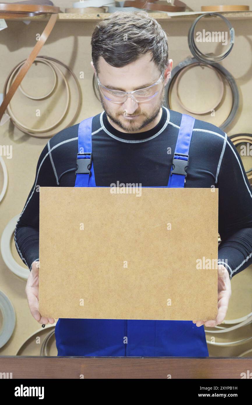 Le fabricant de meubles dans des vêtements spéciaux et des lunettes garde une plaque vide pour le texte sur leur lieu de travail. Fabrication artisanale de meubles Banque D'Images