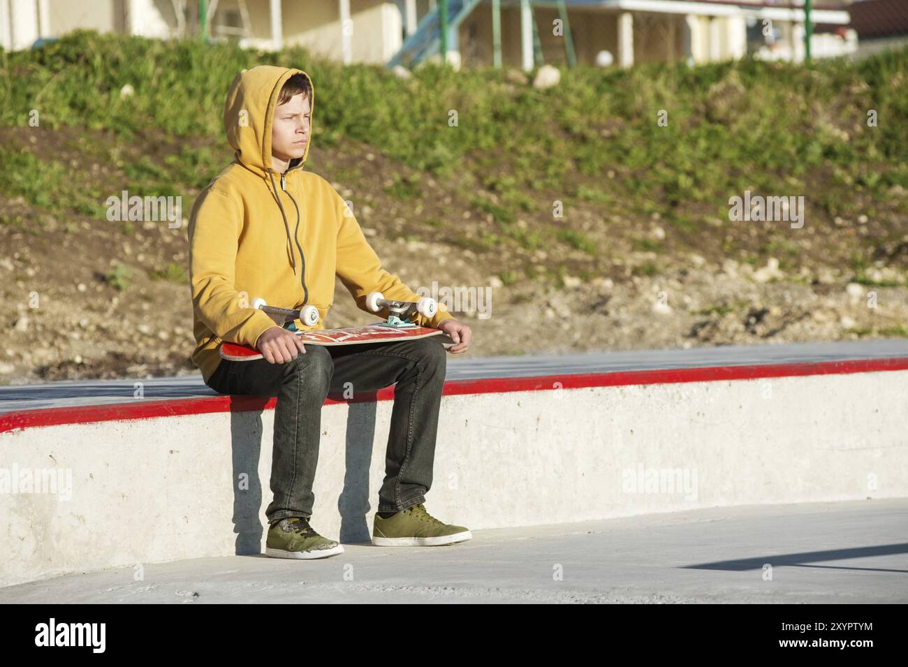 Adolescent vêtu d'un jeans sweat-shirt et de baskets assis dans un skate Park regardant au loin et tenant une planche à roulettes Banque D'Images