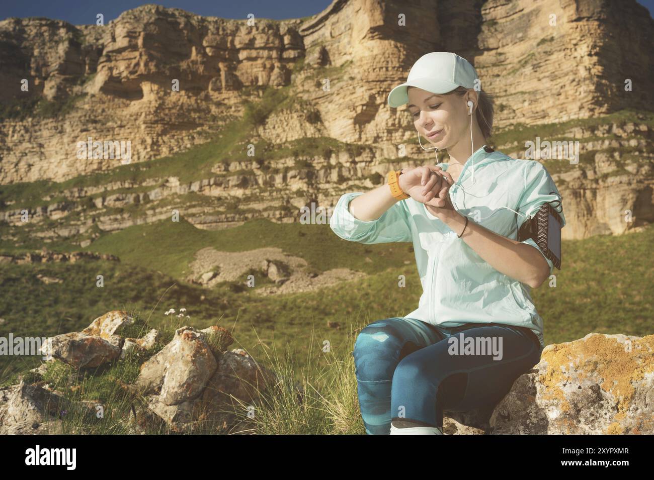 Portrait d'une jeune fille de fitness souriante dans une casquette et des écouteurs vérifiant son horloge intelligente assise sur un rocher à l'extérieur sur un fond de rochers Banque D'Images