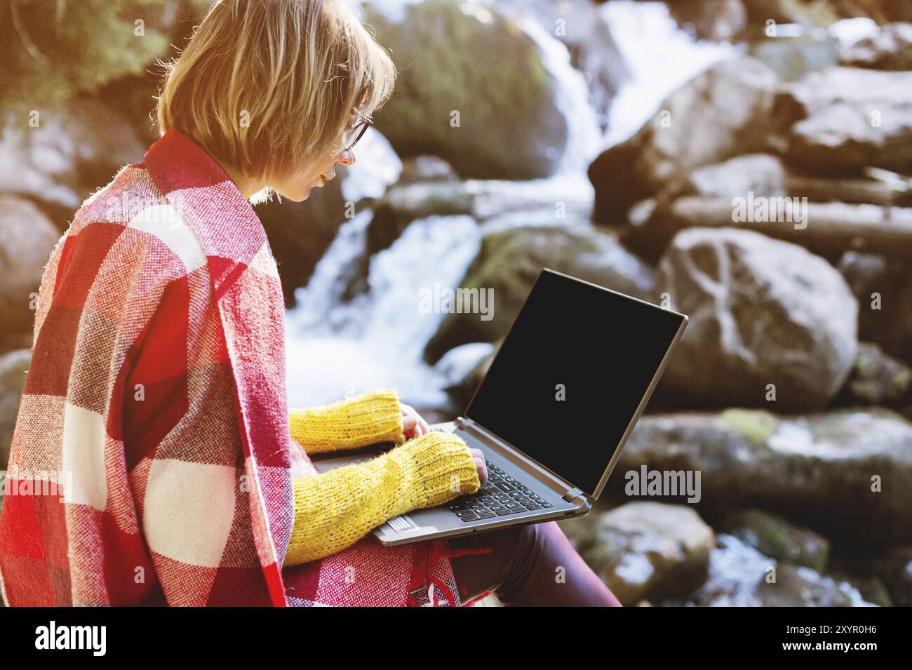 Image maquette d'une fille enveloppée dans un plaid avec un ordinateur portable avec un bureau noir vierge sur ses genoux sur le fond d'une rivière de montagne et lar Banque D'Images