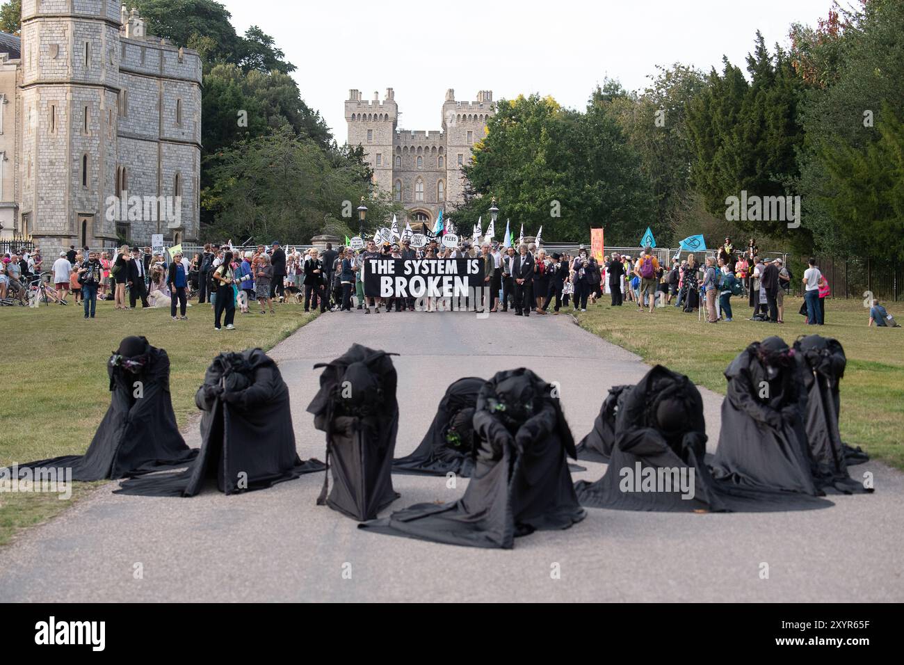 Windsor, Royaume-Uni. 30 août 2024. Extinction Rebellion a organisé une manifestation sur la longue marche avec pour toile de fond le château de Windsor à Windsor, Berkshire ce soir. Les manifestants climatiques de XR portaient des costumes d'affaires et s'habillaient en barons du pétrole, en magnats des médias et en financiers sans visage. Les Oil Slicks, habillés de costumes noirs avec de la peinture de visage noir et blanc, ont réalisé une action théâtrale chorégraphiée dans le but de mettre en évidence le récit XR pour la journée, que le système est brisé. Les rebelles XR campent à Home Park en face du château de Windsor aujourd'hui et pendant le week-end dans le cadre de leur surclassement de trois jours Banque D'Images