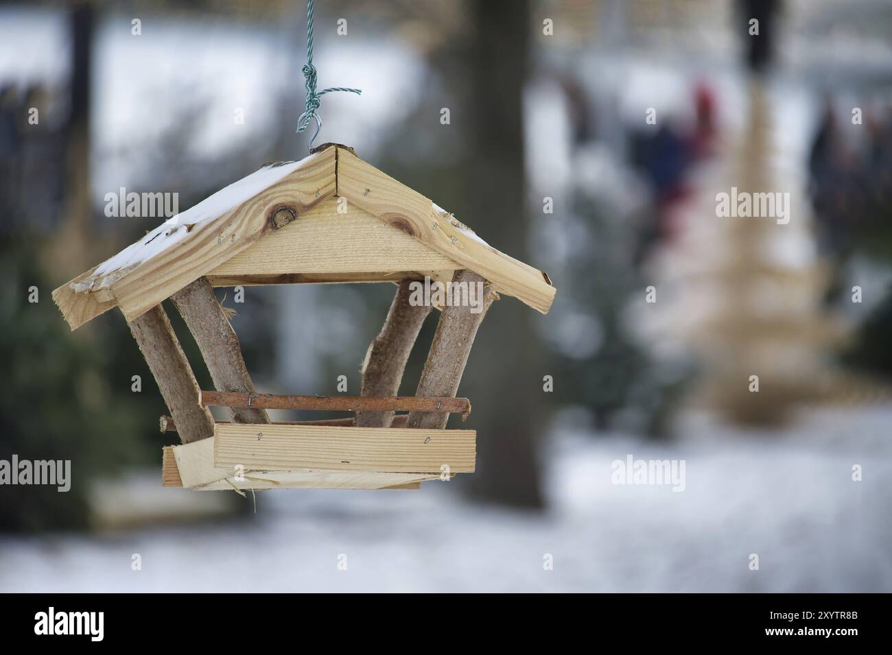 Mangeoire à oiseaux en bois, en forme de petite maison, suspendue à une ficelle dans un environnement enneigé, le fond est flou, contenant des arbres couverts de neige Banque D'Images