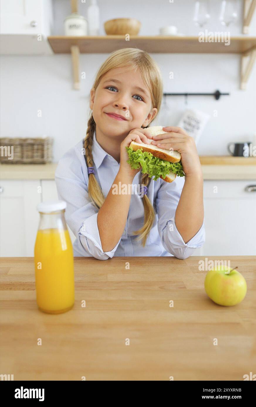 Happy little girl d'âge scolaire, l'enfant blond bénéficiant d''un petit-déjeuner sain eating sandwich et fruits et jus d'orange Boisson assis à bright sunny ki Banque D'Images