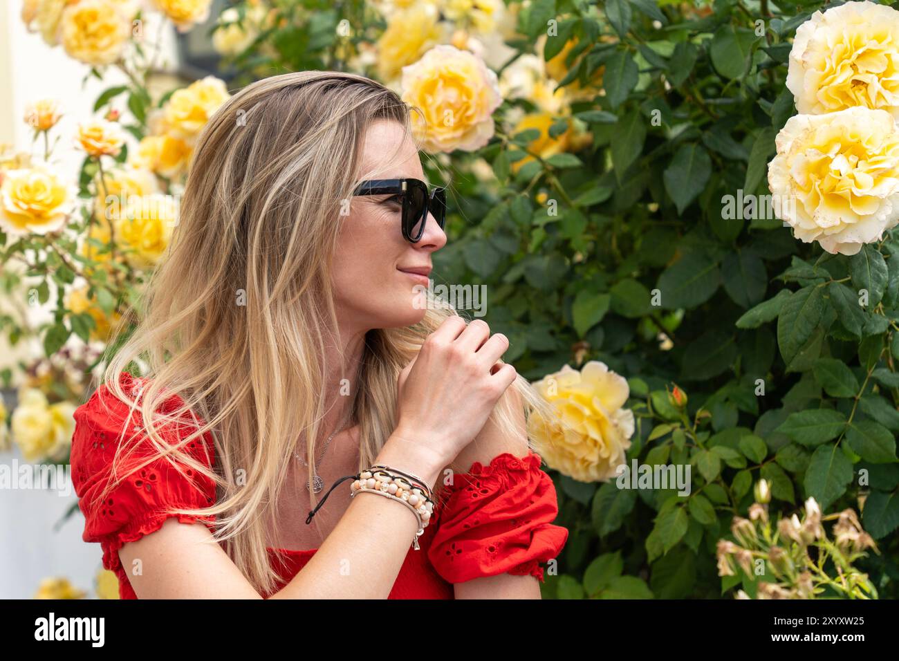 Jeune femme blonde en lunettes de soleil entourée de roses jaunes dans un jardin Banque D'Images