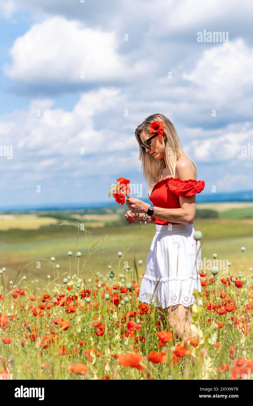 Jeune femme blonde dans des lunettes de soleil avec des coquelicots dans ses cheveux cueillant des fleurs dans un champ de coquelicots rouges. Banque D'Images