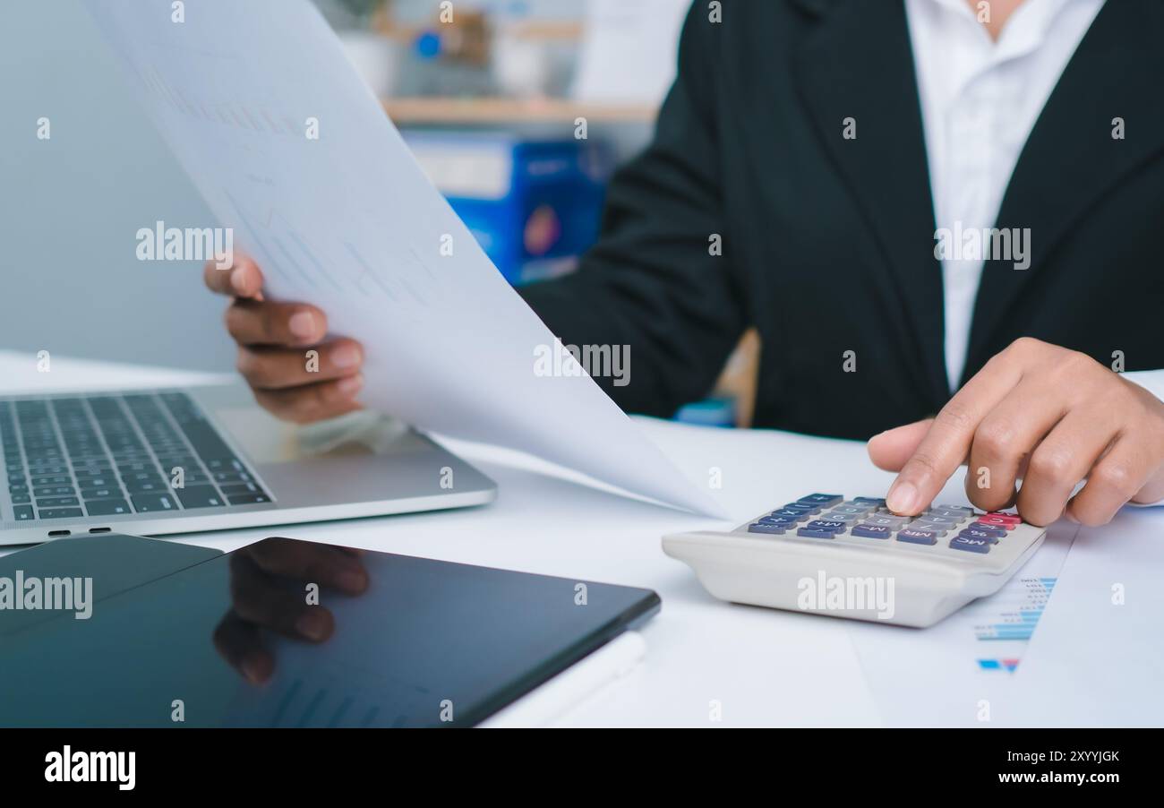 Femme d'affaires en costume noir travaillant sur ordinateur portable, documents papier, tablette et calculatrice, les mains touchent sur tablette au bureau avec fond sombre, Banque D'Images
