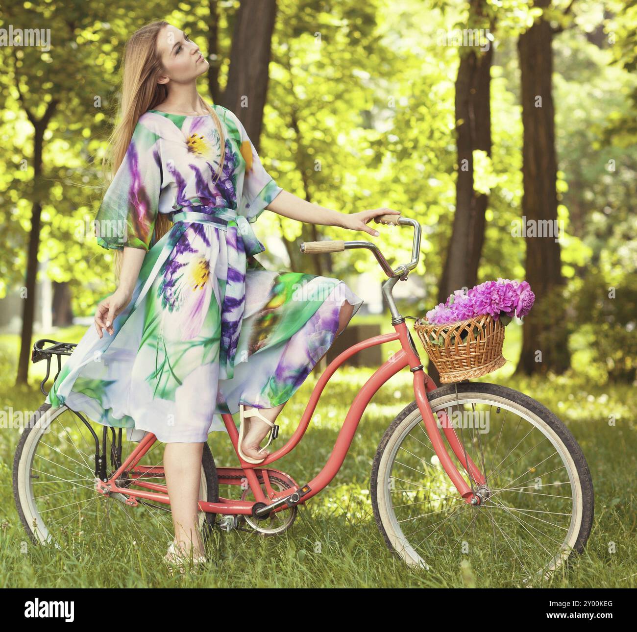 Belle femme blonde portant une belle robe s'amusant dans le parc avec vélo portant un beau panier plein de fleurs de pivoine. Paysage vintage. Joli Banque D'Images