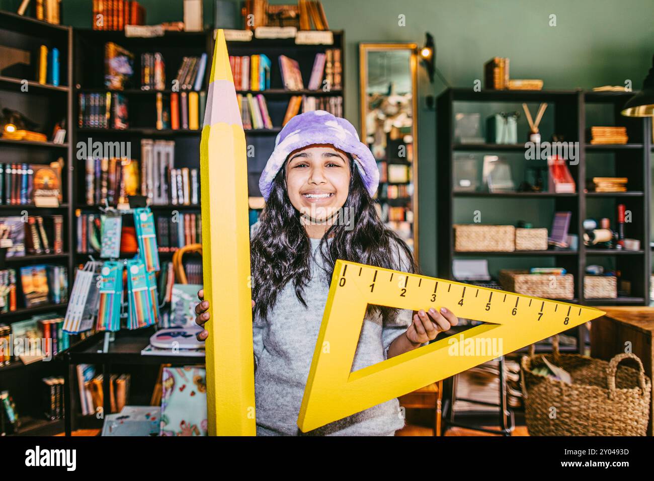 Portrait d'une fille souriante tenant un crayon jouet et placé carré tout en se tenant debout dans la librairie Banque D'Images