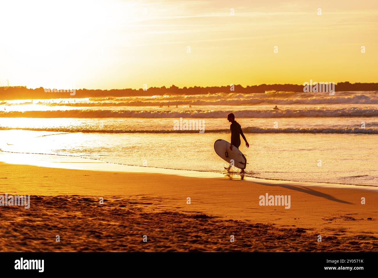 Un homme marche sur la plage avec une planche de surf. Le ciel est orange et l'eau est calme Banque D'Images