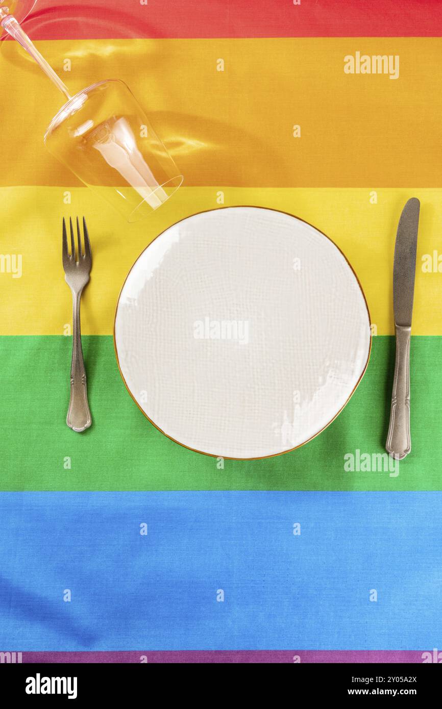 Motif dîner gay. Une assiette avec des couverts et un verre à vin, plan de dessus plat sur la texture du drapeau arc-en-ciel LGBTQ Banque D'Images