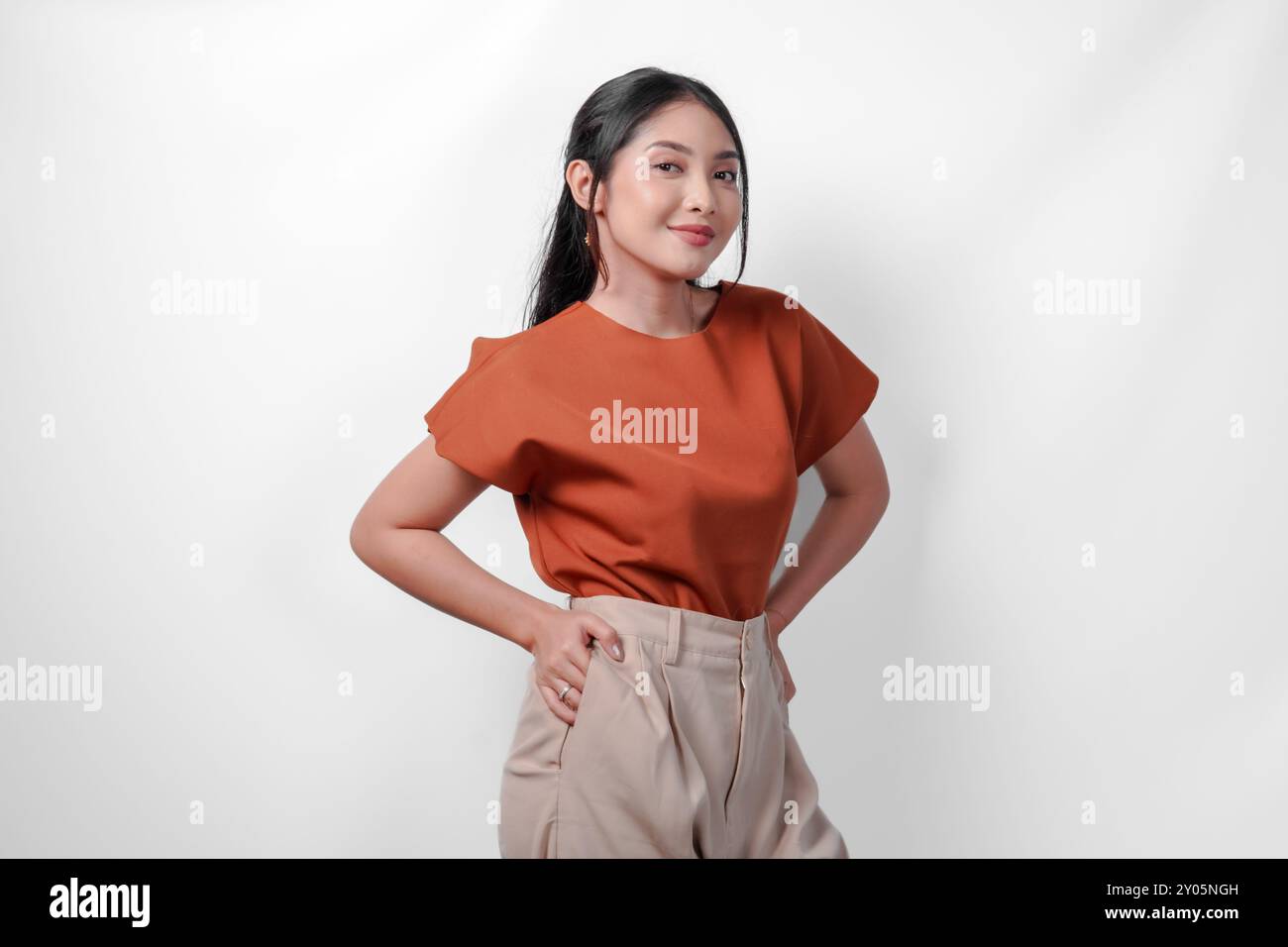 Belle jeune femme asiatique en chemise brune et pantalon taupe plaçant les mains sur la hanche avec une expression confiante, isolée par fond blanc. Banque D'Images