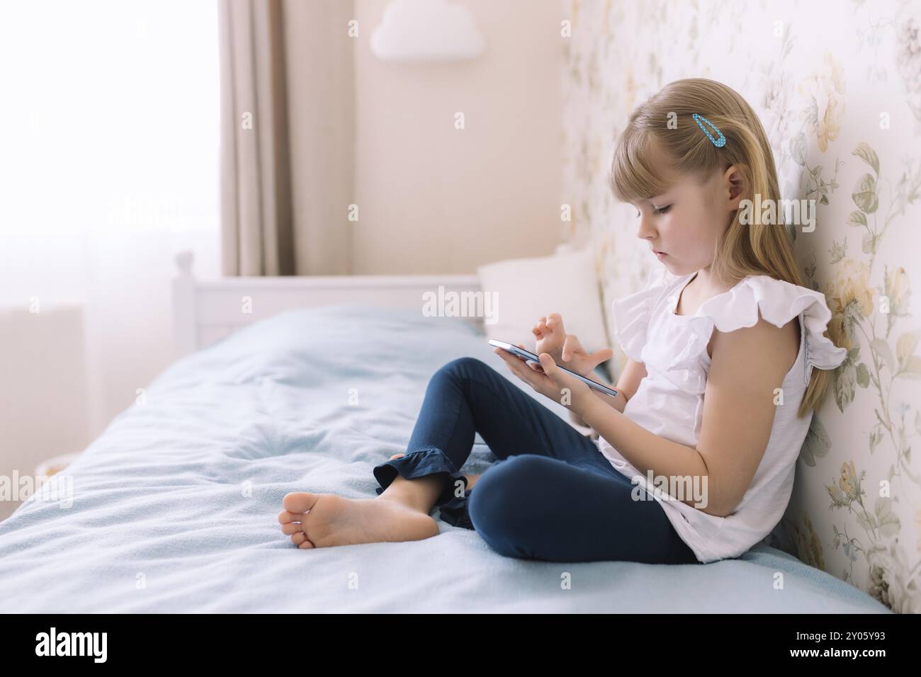 Une petite fille s'assoit sur le lit dans la chambre élégante, tenant le téléphone et lit quelque chose dans le smartphone. Concept de communication Banque D'Images