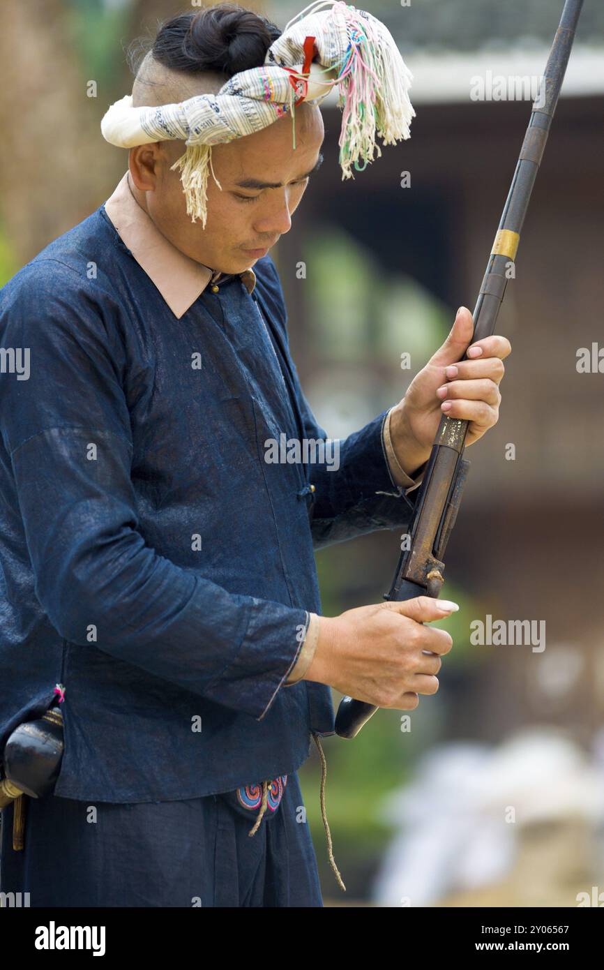 Biasha, Chine, 17 septembre 2007 : homme de la minorité ethnique Miao en vêtements bleus traditionnels, coiffe tressée et chignon aux cheveux longs chargeant une arme à feu à Miao vil Banque D'Images