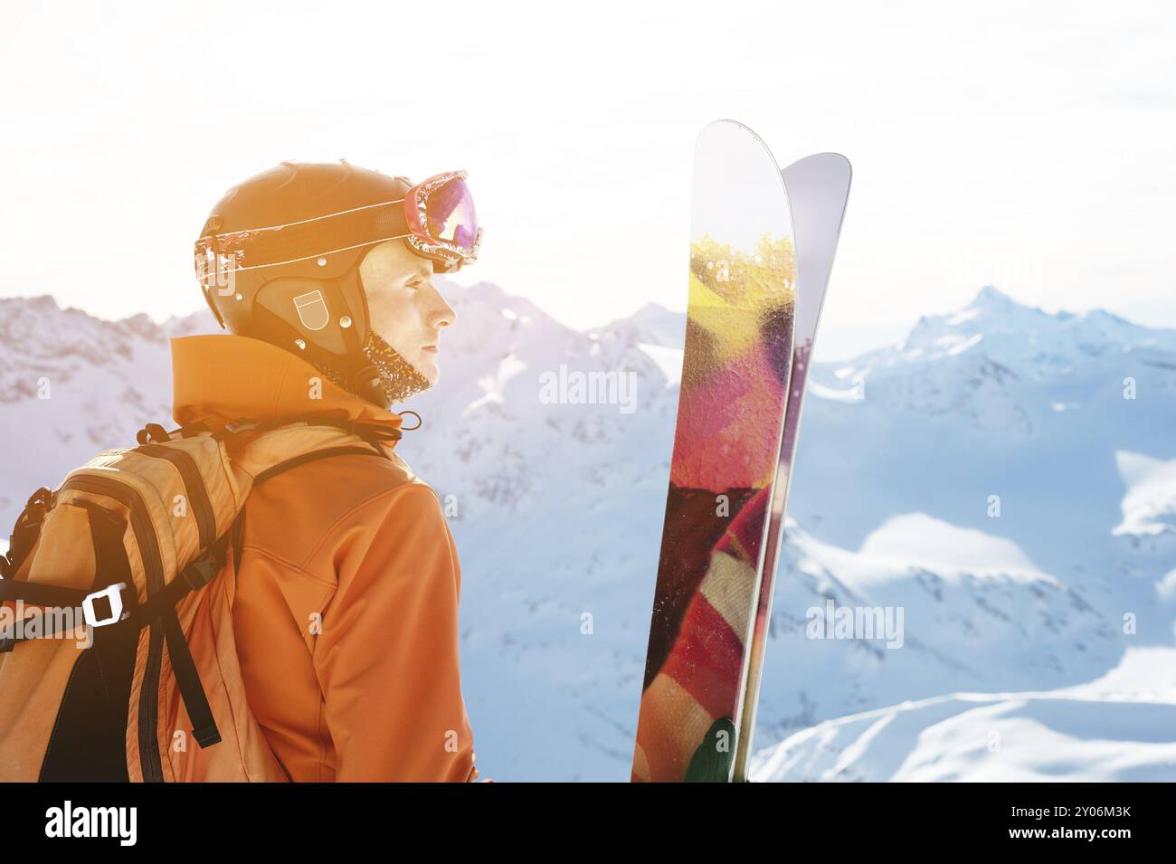 Un skieur dans une combinaison orange avec un sac à dos sur le dos portant un casque et avec des bâtons de ski dans ses mains se tient debout sur un précipice devant un sno Banque D'Images