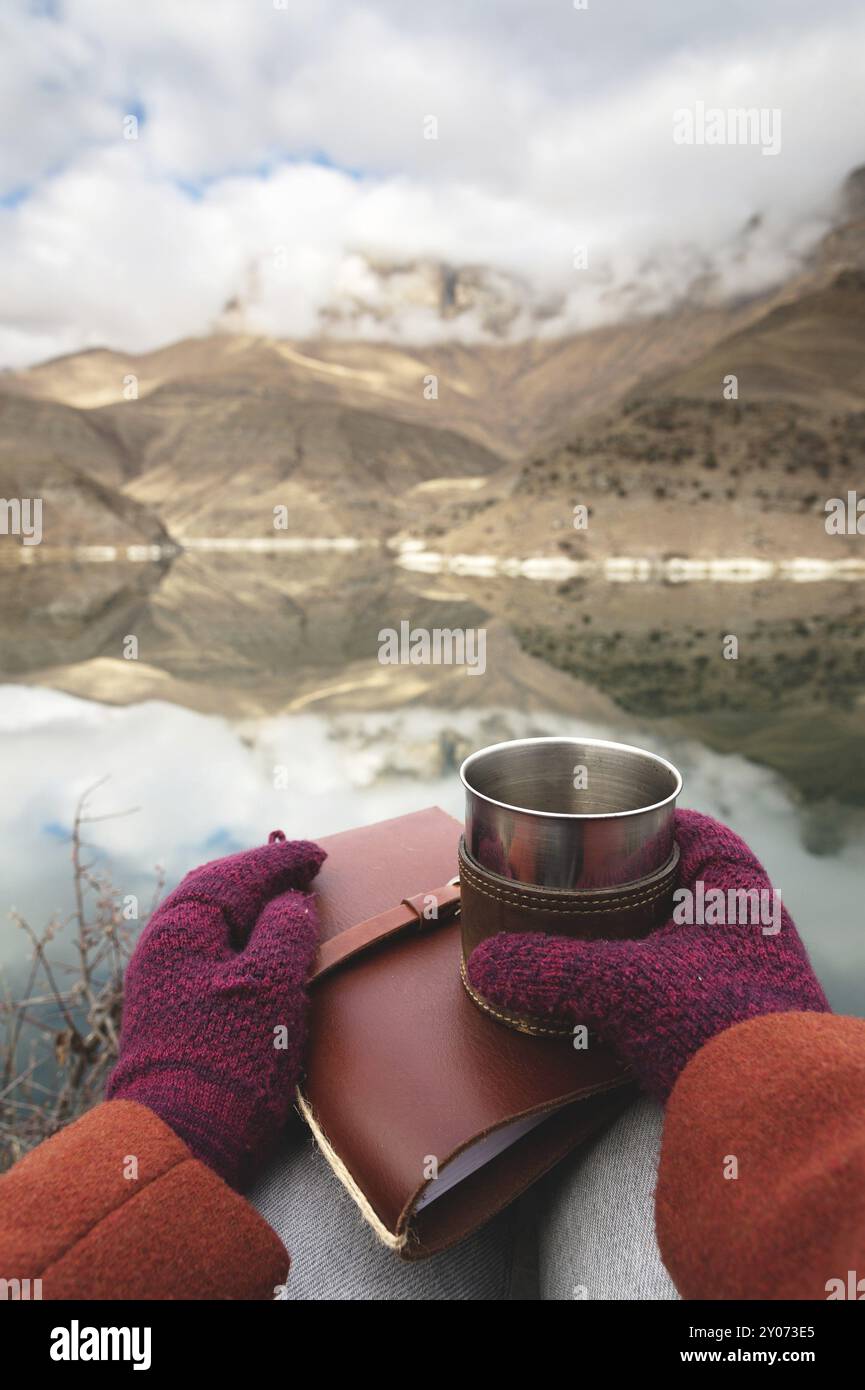 Une vue à la première personne des mains des femmes dans les manteaux et les mitaines tiennent une tasse en métal avec du thé ou du café et un journal de poche en cuir sur la toile de fond o Banque D'Images